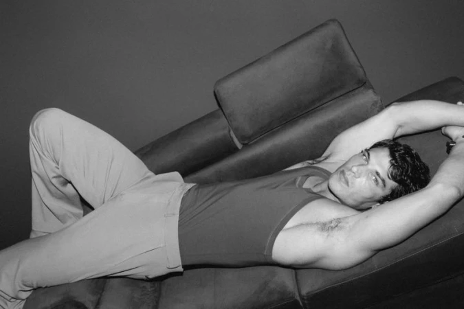 Model Stefano Tomadini reclining on a dark lounge chair wearing a sleeveless tank top and tailored trousers in black and white.