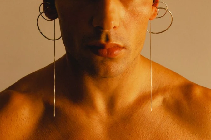 Close-up portrait of model Stefano Tomadini wearing sculptural silver hoop and drop earrings against a warm background.
