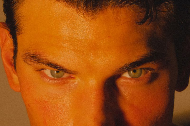 Extreme close up of model Stefano Tomadini with green eyes and dark hair bathed in warm orange and amber lighting.