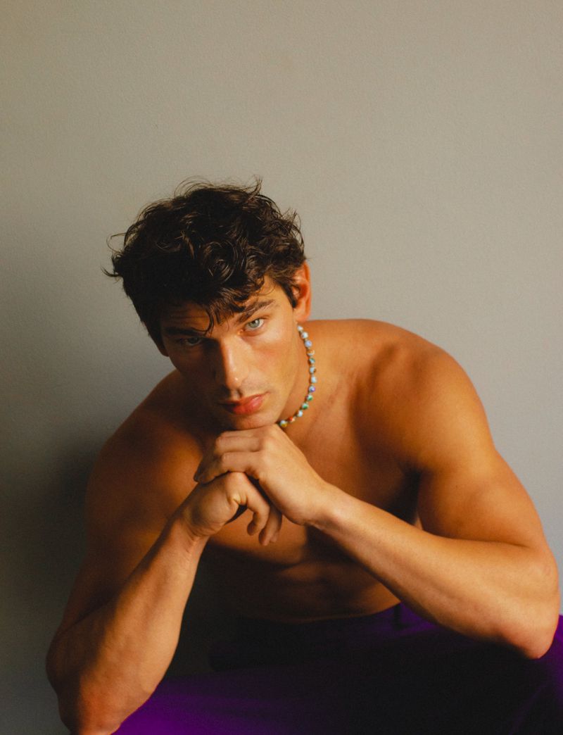 Shirtless man Stefano Tomadini resting chin on clasped hands while wearing a beaded necklace against a grey backdrop.