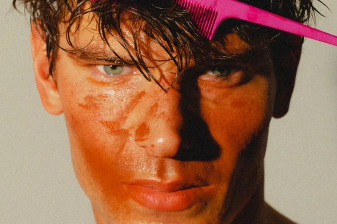 Close-up portrait of a male model with wet hair holding a bright pink comb in his hair against a plain neutral background.