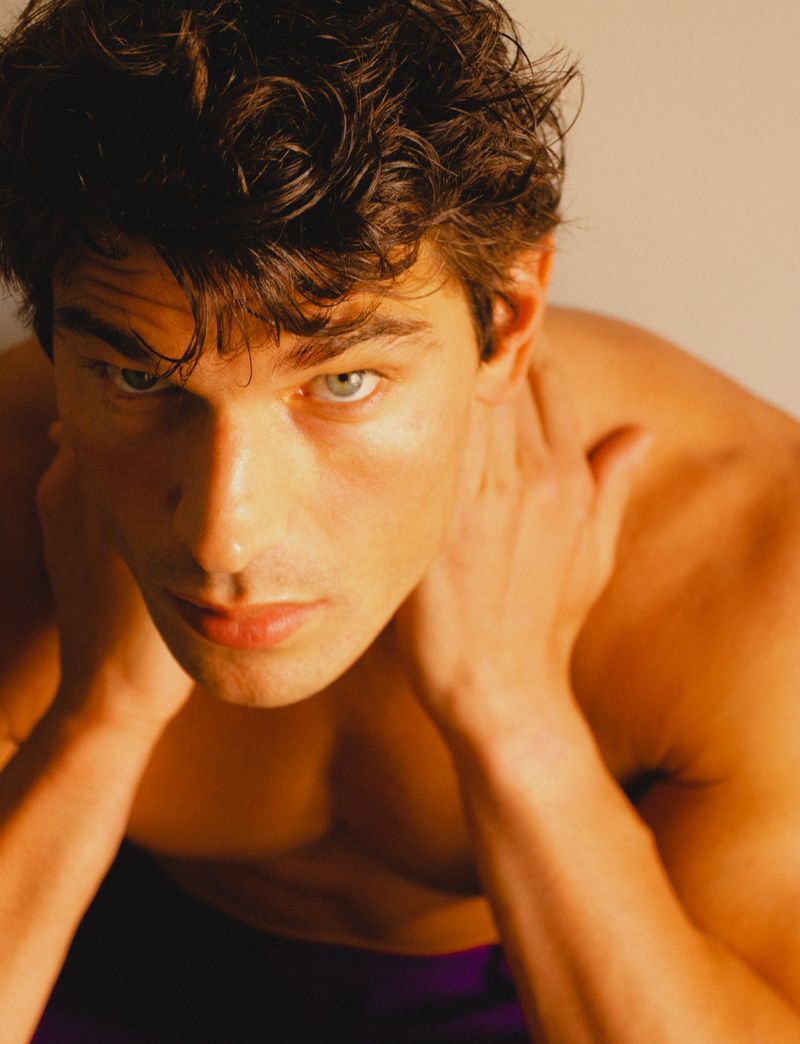 Close-up portrait of model Stefano Tomadini posing shirtless with hands behind his neck under warm lighting.