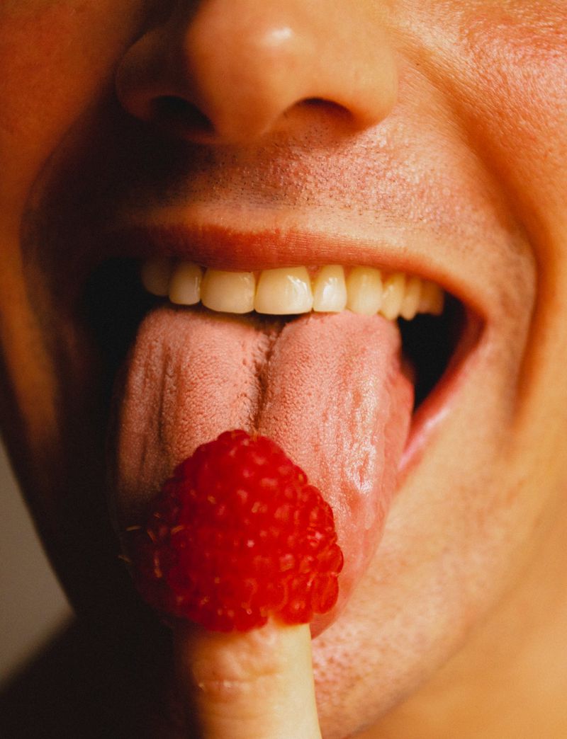 Extreme close-up of a model sticking his tongue out to touch a vibrant red raspberry balanced on his fingertip.