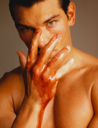 Close-up portrait of a shirtless man with slicked-back hair holding a hand covered in glossy reddish liquid over his face.