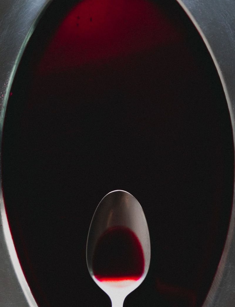 A metal spoon resting at the bottom of an oval frame filled with deep crimson liquid against a dark backdrop.
