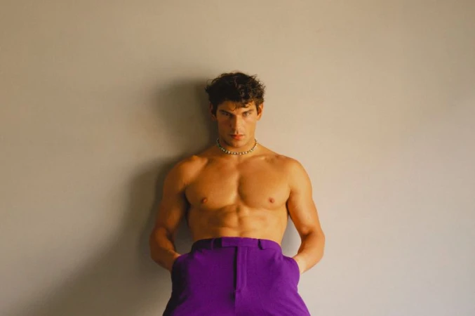Model Stefano Tomadini posing shirtless in bright purple tailored trousers and a beaded choker necklace against a neutral wall.