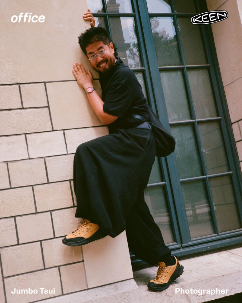 Man smiling while clinging to an exterior stone wall ledge wearing a black outfit and mustard yellow KEEN sneakers.