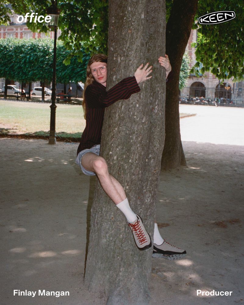 Finlay Mangan clinging to a tree trunk wearing shorts and KEEN sneakers in an outdoor park setting.