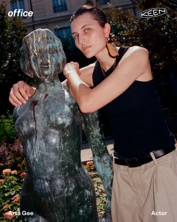 Model Arta Gee wearing a black tank top and beige trousers while leaning against a weathered bronze statue in a garden.