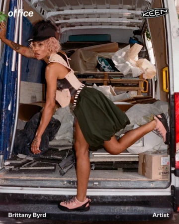 Brittany Byrd posing inside the open rear of a cargo van wearing a tan vest, olive balloon skirt, and pink KEEN shoes.