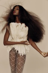 Black woman with long flowing dark hair wearing a white ruffled top and patterned tights against a light background.
