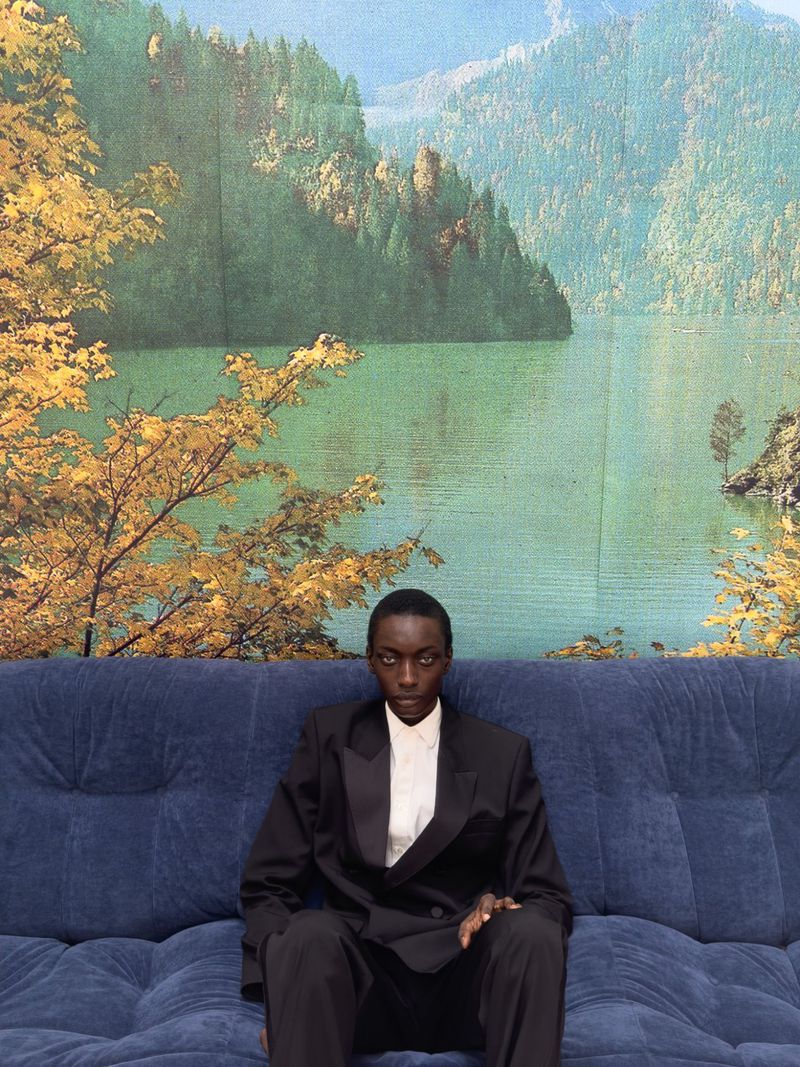 Adult in black suit and white dress shirt sits on a navy couch in front of a mountain landscape painting backdrop. Portrait orientation.