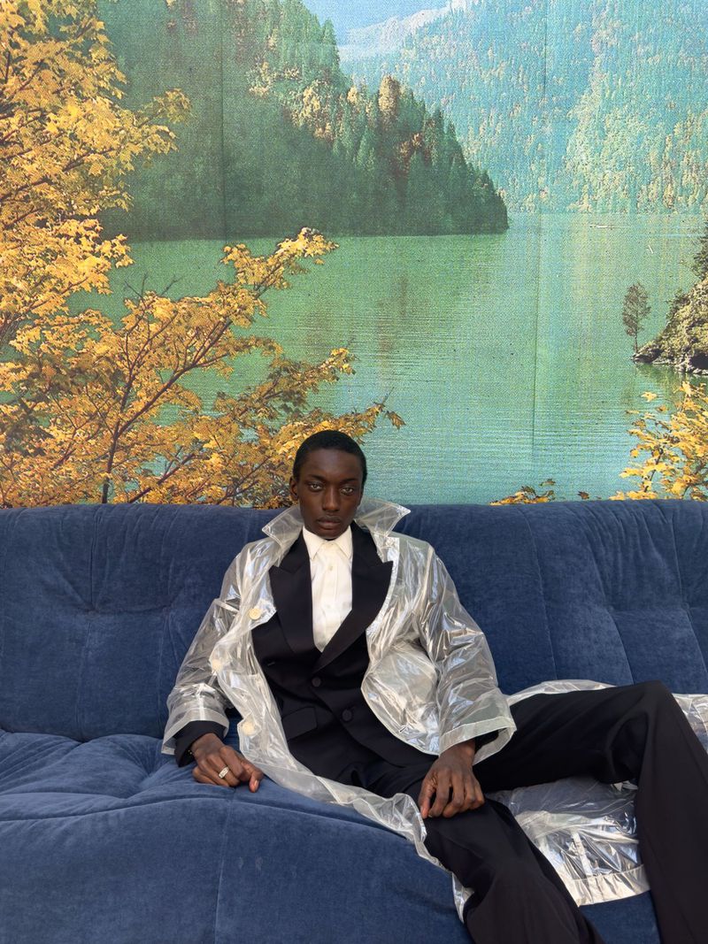A single young adult man lounges on a blue sofa wearing a black suit and a clear plastic coat in front of a nature-scene backdrop.