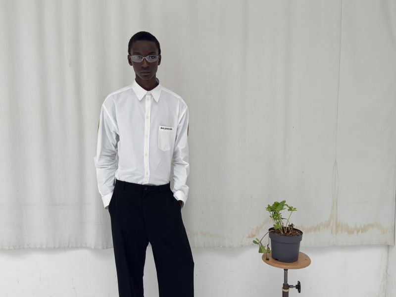 Adult male in white button-down shirt and dark pants poses against a white backdrop, near a plant stand with a potted plant.