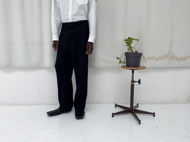 Man in white shirt and black dress pants poses next to a green potted plant in a bright indoor studio setting with neutral colors.