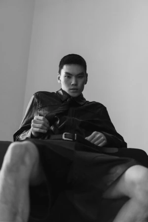 Low-angle black and white portrait of model Buyant Munkhbat seated in a leather jacket and dark shorts.