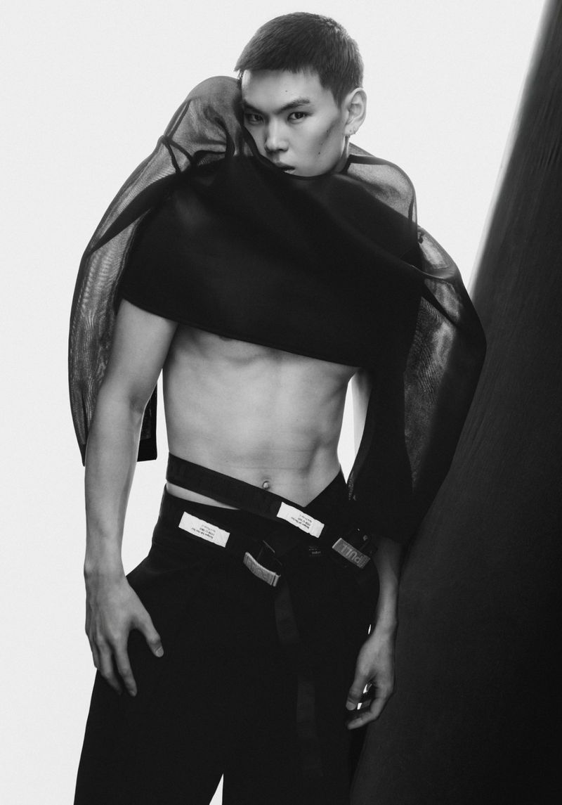Model Buyant Munkhbat posing shirtless with a sheer black fabric wrap and black trousers in a black and white studio shoot.