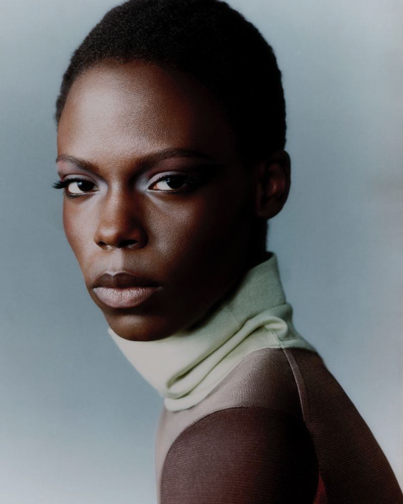 Close-up portrait of model Agel Akol with short hair wearing a pale high-neck collar against a soft grey background.