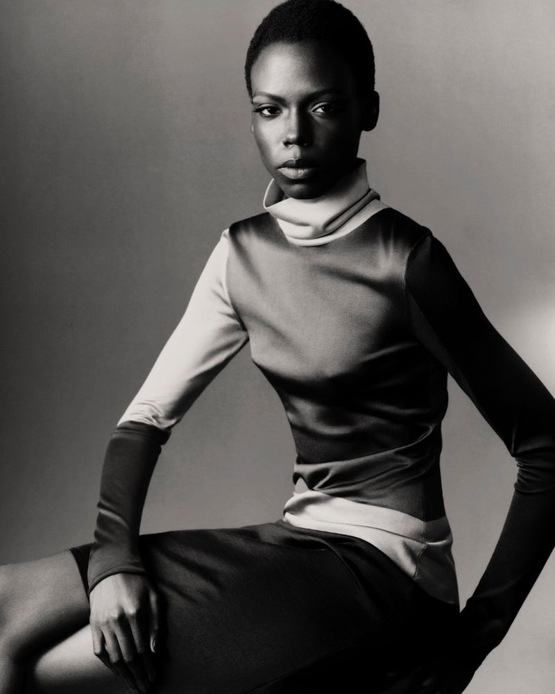 Model Agel Akol seated in a high-neck long-sleeve top and skirt in monochrome against a studio backdrop.