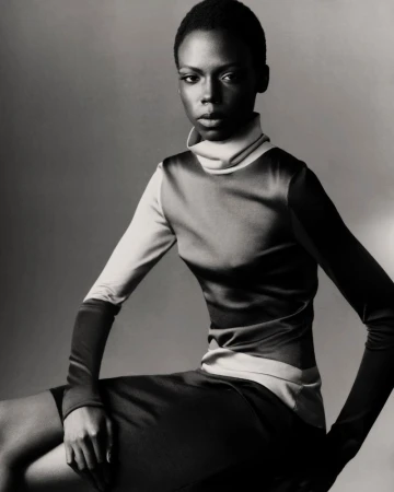 Model Agel Akol seated in a high-neck long-sleeve top and skirt in monochrome against a studio backdrop.