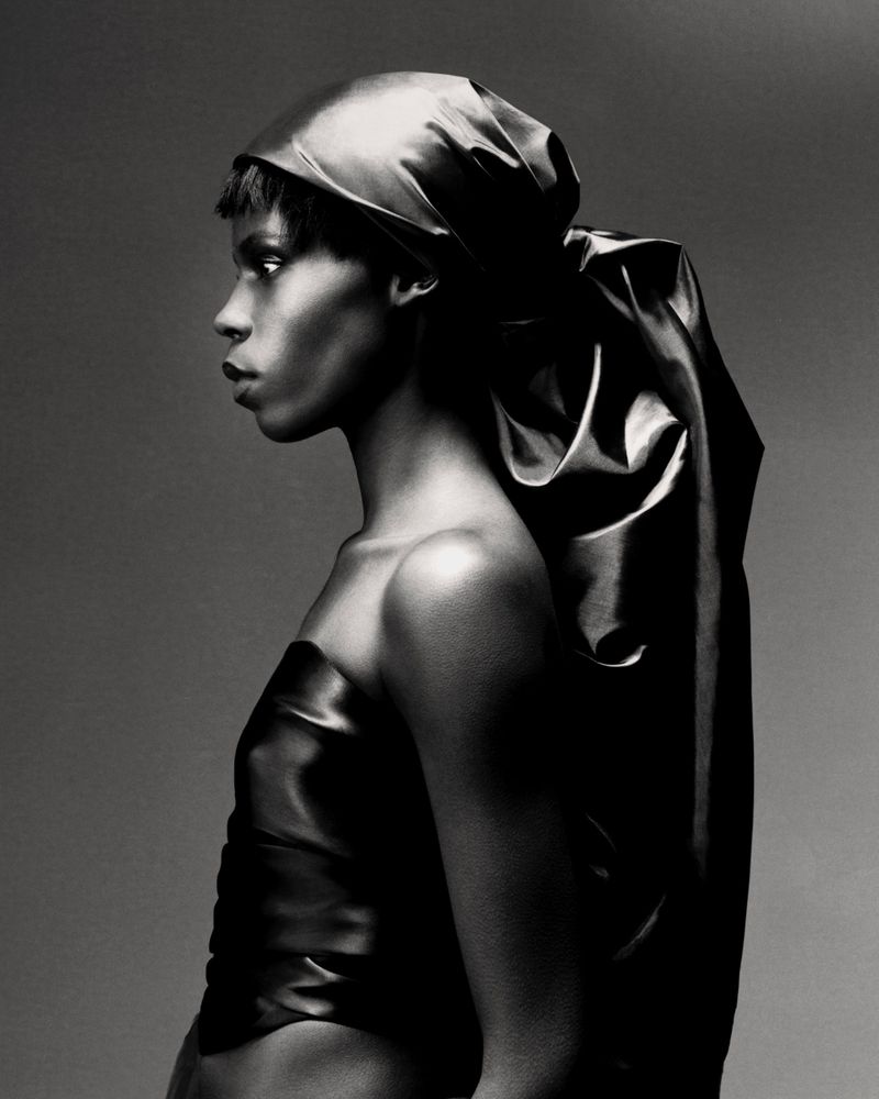 Model Agel Akol in profile wearing a glossy draped satin headscarf and matching strapless top in a black and white studio portrait.