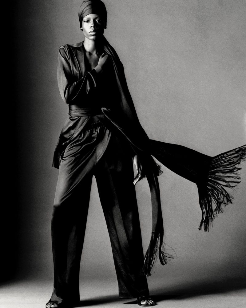 Model Agel Akol in a monochrome draped outfit with a matching headwrap and flowing fringed scarf against a grey background.