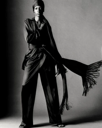 Model Agel Akol in a monochrome draped outfit with a matching headwrap and flowing fringed scarf against a grey background.