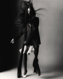Monochrome fashion portrait of Agel Akol wearing a black fringe-accented coat with flowing hair in motion against a studio backdrop.