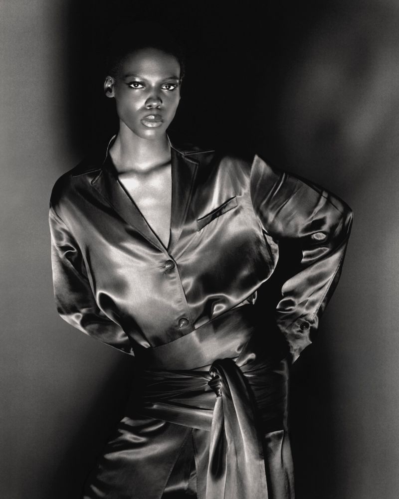 Model Agel Akol wearing a glossy satin button-down shirt and tied waist wrap in a black and white studio portrait.