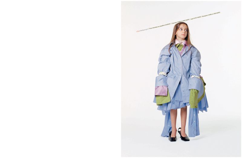 Young girl standing in oversized layered blue shirts, mismatched collars, and oversized black heels against a white backdrop.