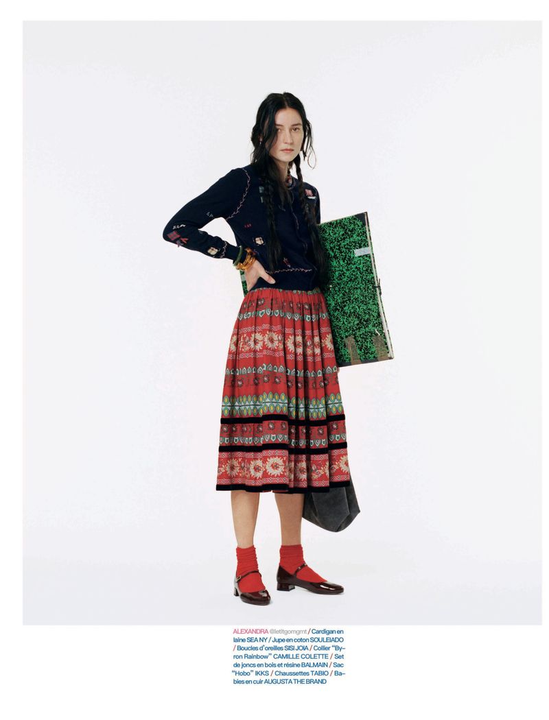 Model Alexandra Zalivako standing in a navy cardigan and red patterned pleated skirt with a green case against a white background