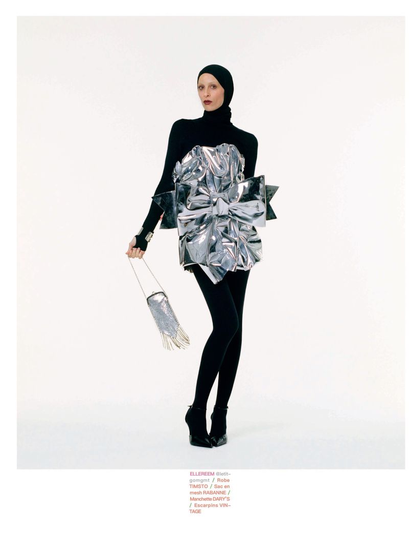 Model Elle Reem posing in a black balaclava jumpsuit and an oversized metallic silver bow dress against a white seamless background.