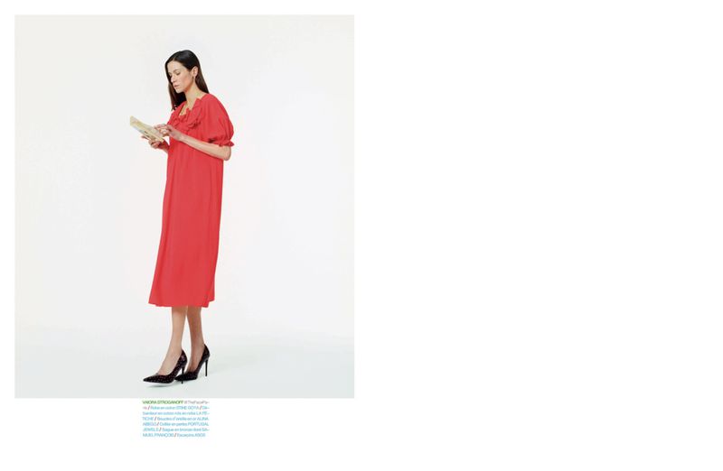 Model Vaiora Stroganoff stands in a bright red midi dress and black pointed heels holding a booklet against a white studio background.