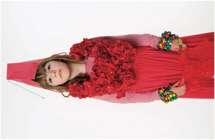 Young girl in a red dress with a red floral chest piece, colorful bead bracelets, and a red paper headpiece against a white background.