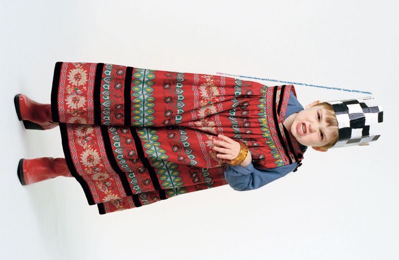 A young child wearing a red patterned cape dress, blue shirt, red boots, and a checkered paper crown against a white backdrop.