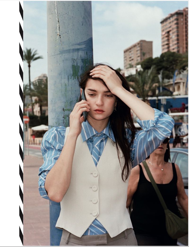 Model leaning against a post in a blue striped shirt and white vest making a phone call outdoors in Benidorm