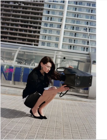 A woman crouching on a paved walkway searching inside an overturned black tote bag in front of modern apartment buildings.