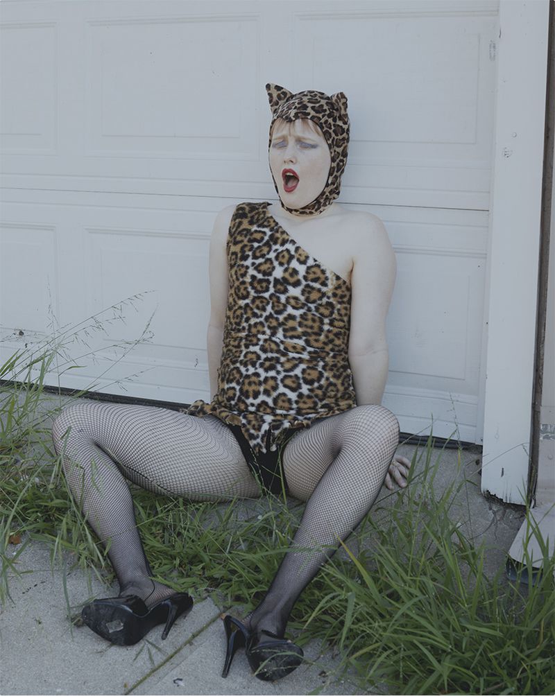 Bobbi Menuez seated against a white garage door wearing a leopard-print cat-ear hood, matching tunic, fishnet tights, and heels.