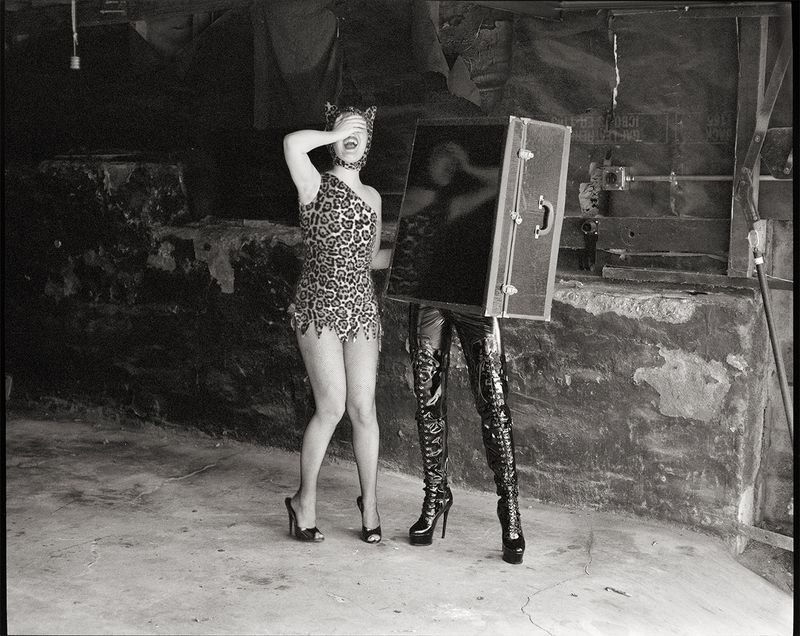 Bobbi Salvör Menuez in a leopard-print suit next to a figure wearing high-heeled vinyl boots inside a black trunk.