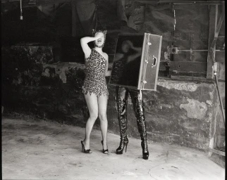 Bobbi Salvör Menuez in a leopard-print suit next to a figure wearing high-heeled vinyl boots inside a black trunk.