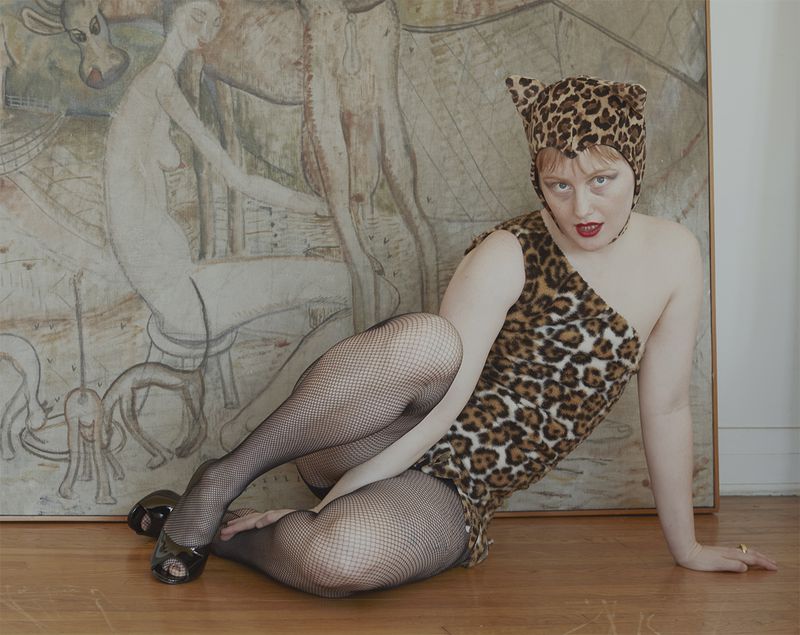 Bobbi Salvör Menuez in a leopard-print one-shoulder romper, matching cat-ear hood, and fishnet tights reclining on a wood floor.