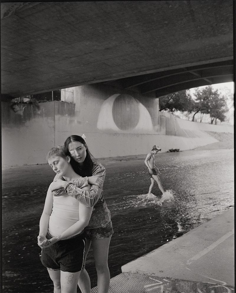Black and white photo of two people embracing under a concrete bridge with a person wading in shallow water behind them.