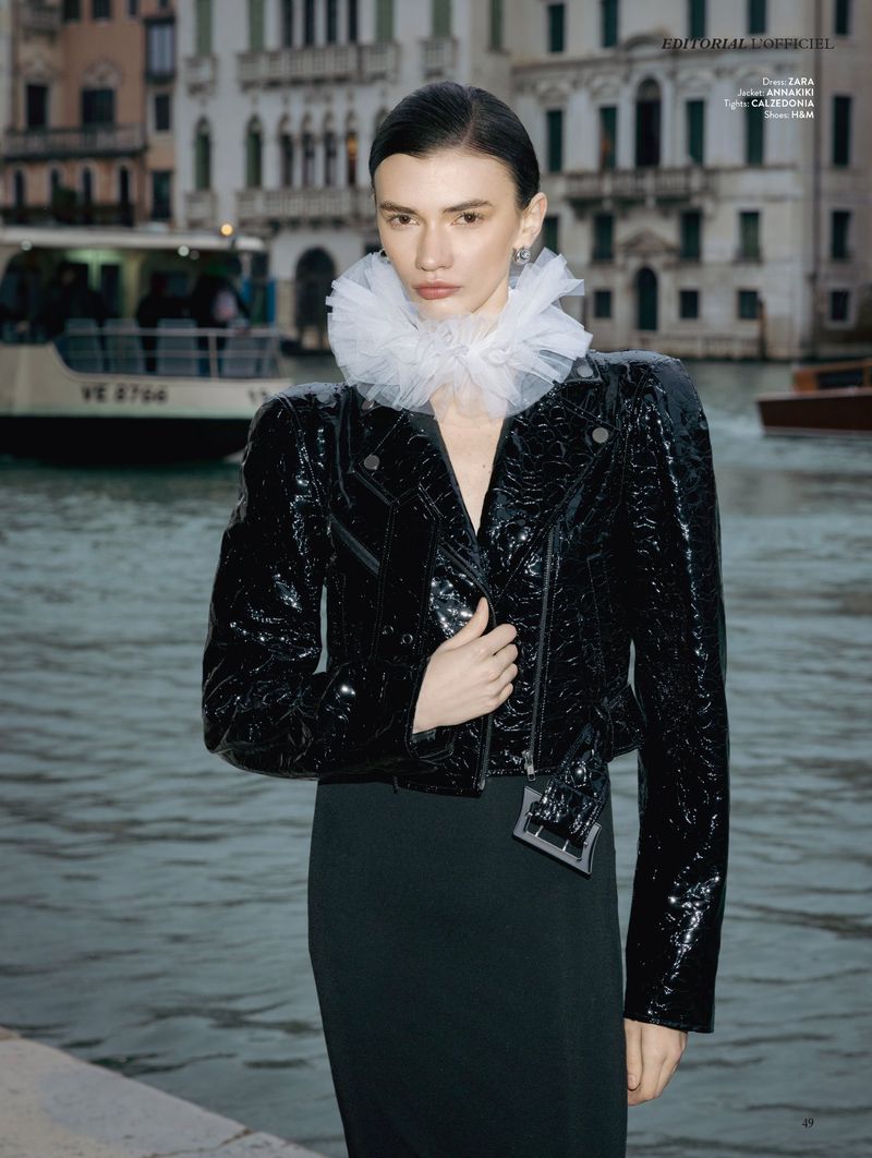 Model Ksenia Yatuvshenko wearing a glossy black jacket and white ruffled tulle collar by a Venice canal.