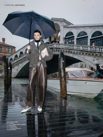Model Ksenia Yatuvshenko holding a black umbrella on a wet wooden dock in Venice with the Rialto Bridge in the background.