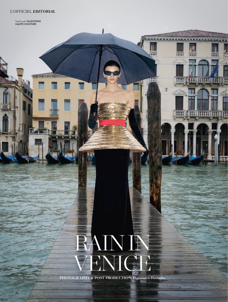 Model in a gold strapless gown holding a black umbrella on a wet wooden dock along the Grand Canal in Venice.