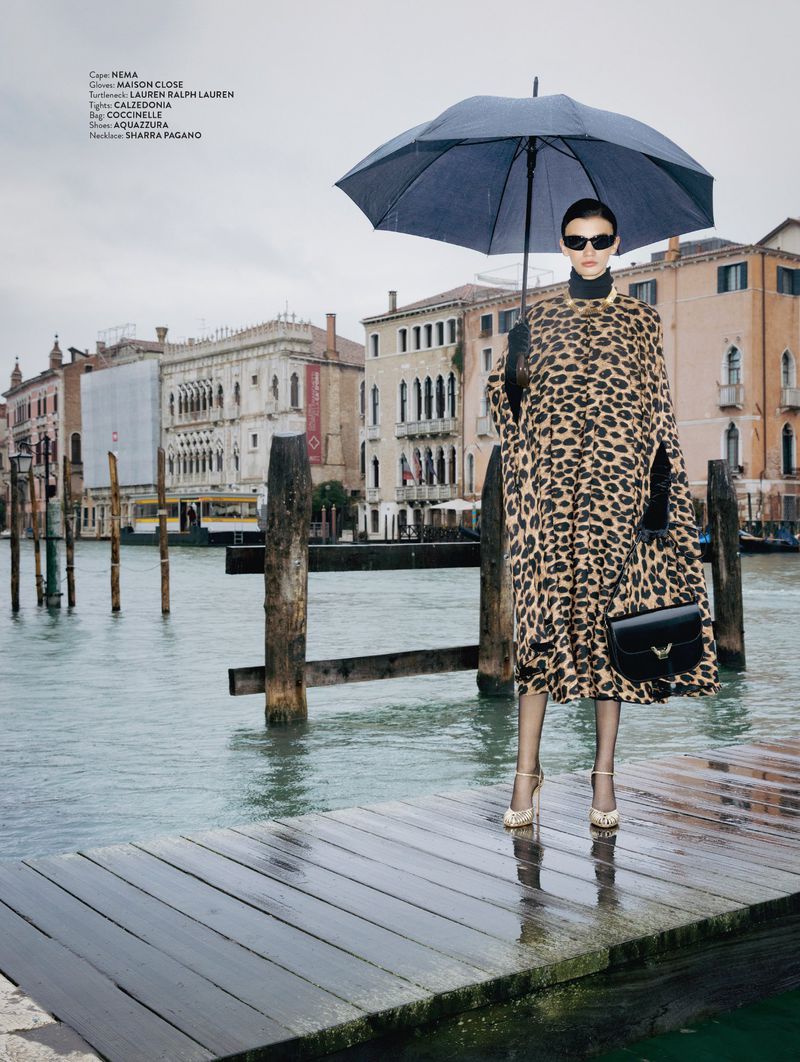 Model Ksenia Yatuvshenko holds a black umbrella on a wooden dock by a Venice canal, wearing a leopard-print cape.