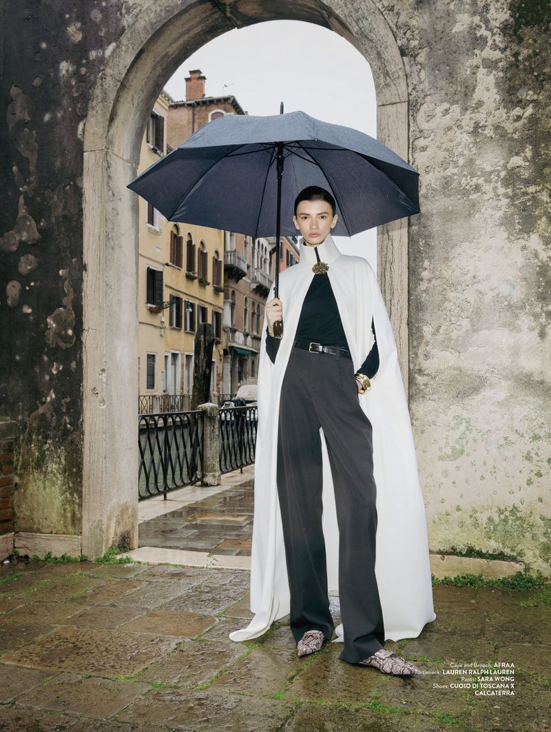 Model Ksenia Yatuvshenko poses in a long white cape holding a black umbrella within a weathered stone arch in Venice.