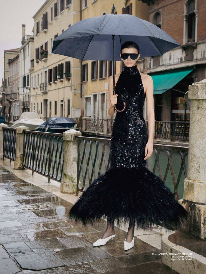 Model wearing a black sequin gown with a feathered hem and dark sunglasses while holding an umbrella beside a canal.