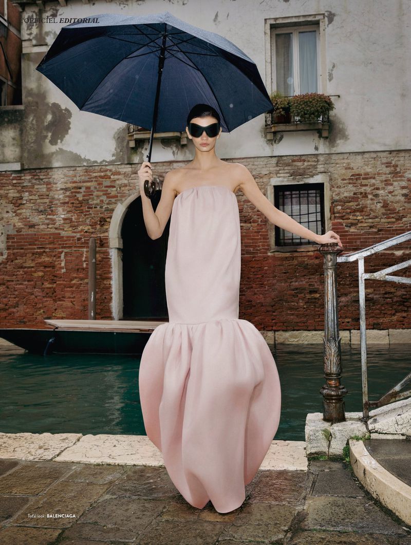 Model wearing a strapless blush pink sculptural gown and futuristic black sunglasses while holding a black umbrella by a canal in Venice.