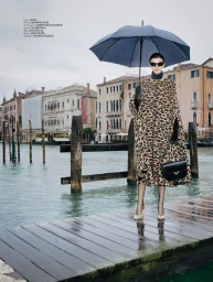 Model Ksenia Yatuvshenko holds a black umbrella on a wooden dock by a Venice canal, wearing a leopard-print cape.